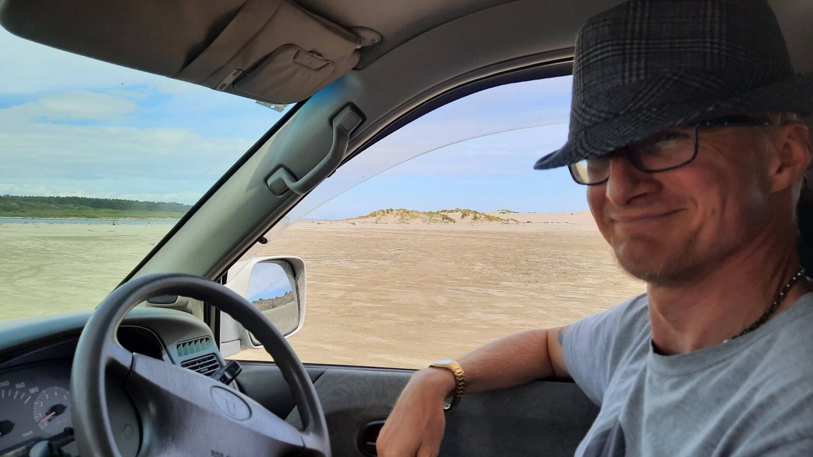 Happy smiling man sits in driver's seat with arm propped on car window opening. A sandy beach is seen out the window.