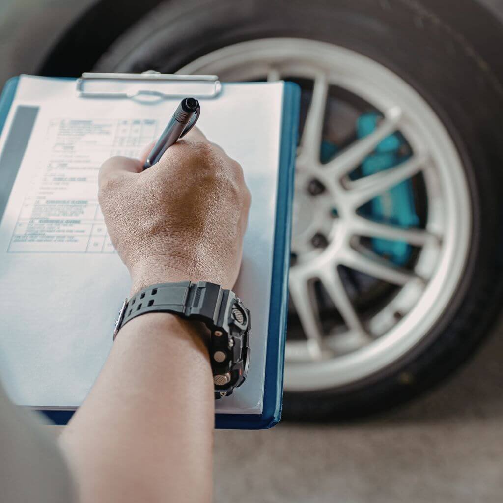 Man holds clipboard and pen in the foreground, a car wheel is seen in the background.